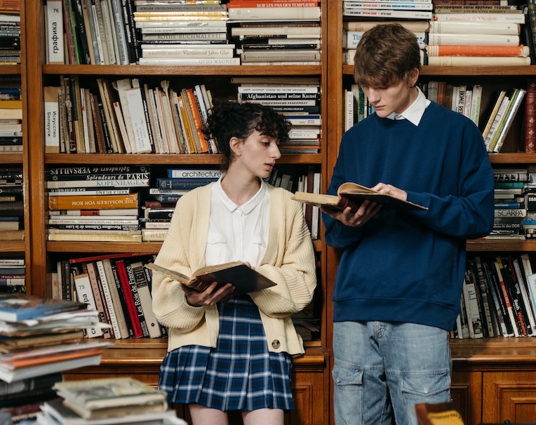 Two students standing next to each other and reading books in a library.